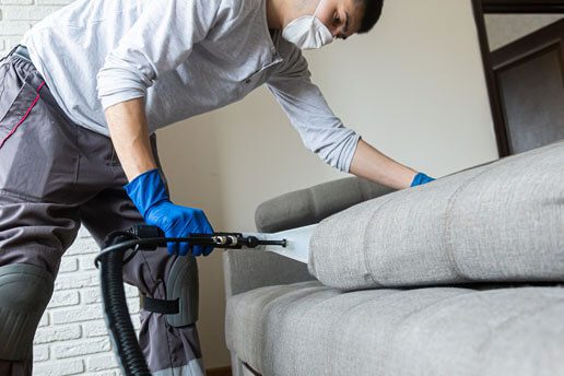 A man in blue gloves is using a vacuum on a couch.